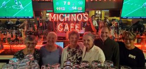 Ron Michno (second from right) in 2025 at Michno’s Café in Redford Township, with his wife of 52 years, She She Michno (far right). Also pictured, left to right, are Mary Ann, Theresa, Aurelia, and Loretta, all daughters of John W. and Stella L. (Michno) ZaydelThe original, bright neon Michno’s Café lighting from the Central Street Michno’s Café shines on behind the bar, paying homage to the enduring Michno’s legacy.